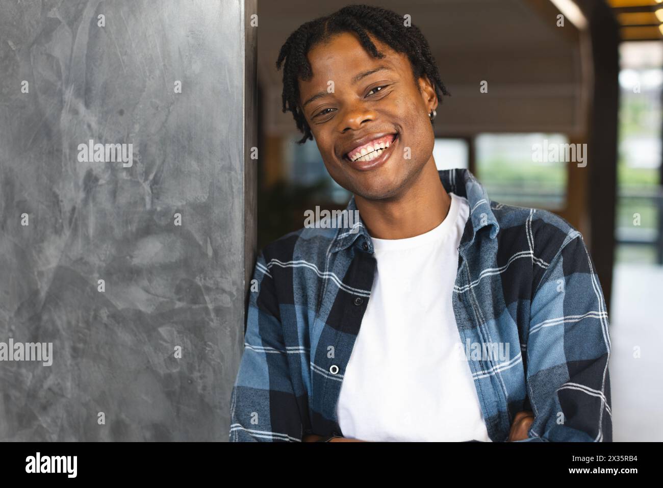 Afroamerikaner in kariertem Hemd lächelt, lehnt sich an eine Wand in einem modernen Büro Stockfoto