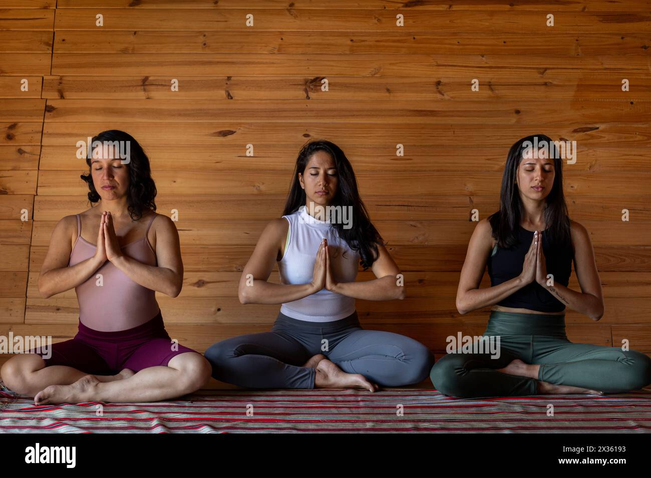 Vorderansicht von drei jungen lateinamerikanischen Frauen, die gemeinsam im Yoga-Unterricht meditieren. Hintergrund der Holzlatten. Konzept von Wellness und Yoga. Stockfoto