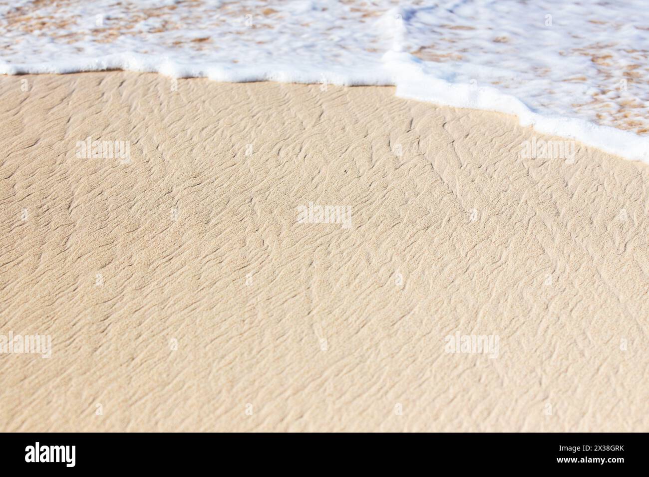 Feuchter weißer Sand und Küstenwasser mit weißem Schaum an einem sonnigen Tag. Natürliches Hintergrundfoto mit selektivem Weichfokus Stockfoto