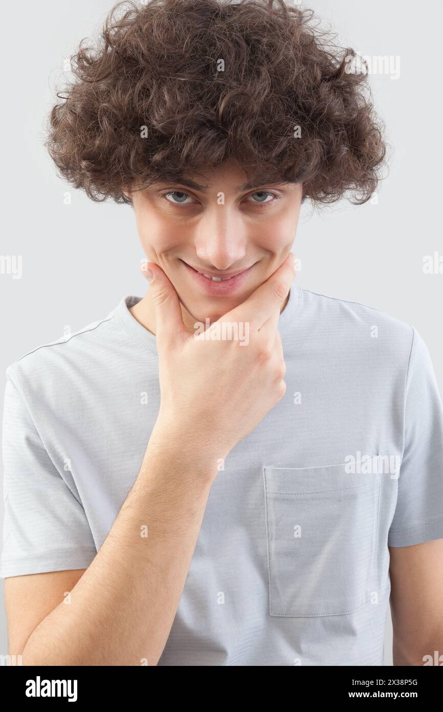 Ein junger Mann mit lockigen Haaren sieht lächelnd in die Kamera, mit seinen blauen Augen, den Händen im Gesicht, einem Porträt eines Mannes mit T-Shirts Stockfoto