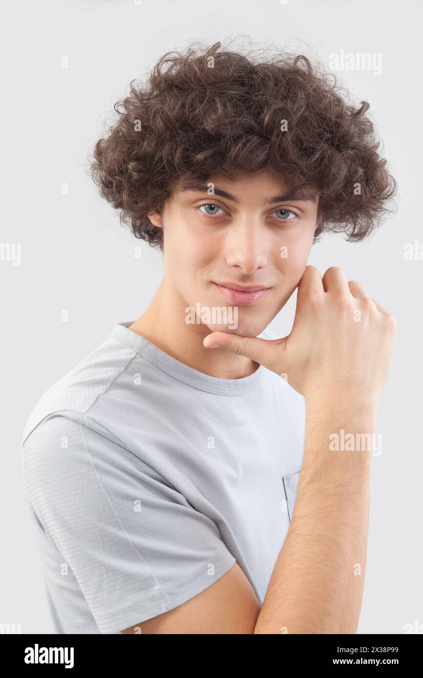 Ein junger Mann mit lockigen Haaren sieht lächelnd in die Kamera, mit seinen blauen Augen, den Händen im Gesicht, einem Porträt eines Mannes mit T-Shirts Stockfoto