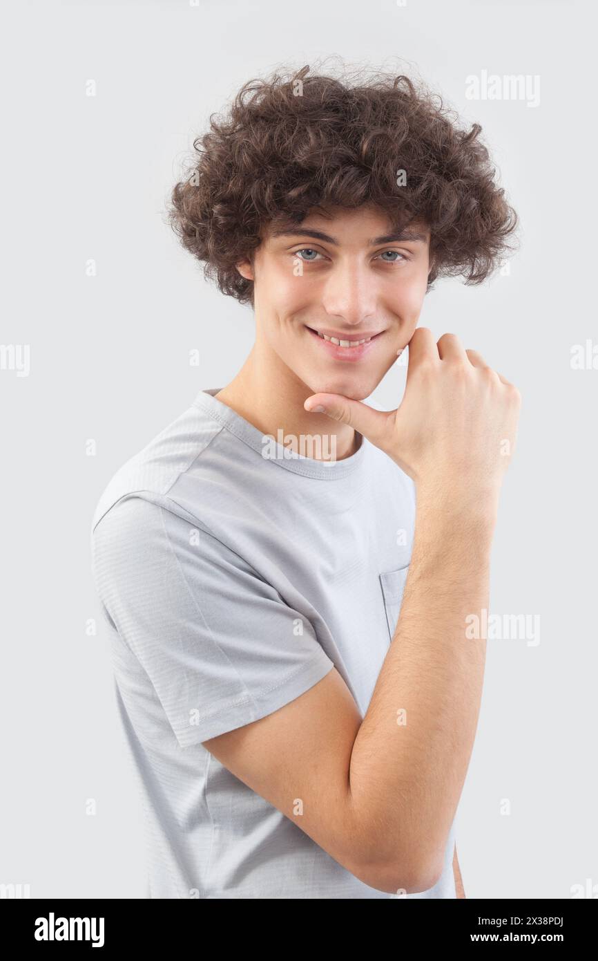 Ein junger Mann mit lockigen Haaren sieht lächelnd in die Kamera, mit seinen blauen Augen, den Händen im Gesicht, einem Porträt eines Mannes mit T-Shirts Stockfoto