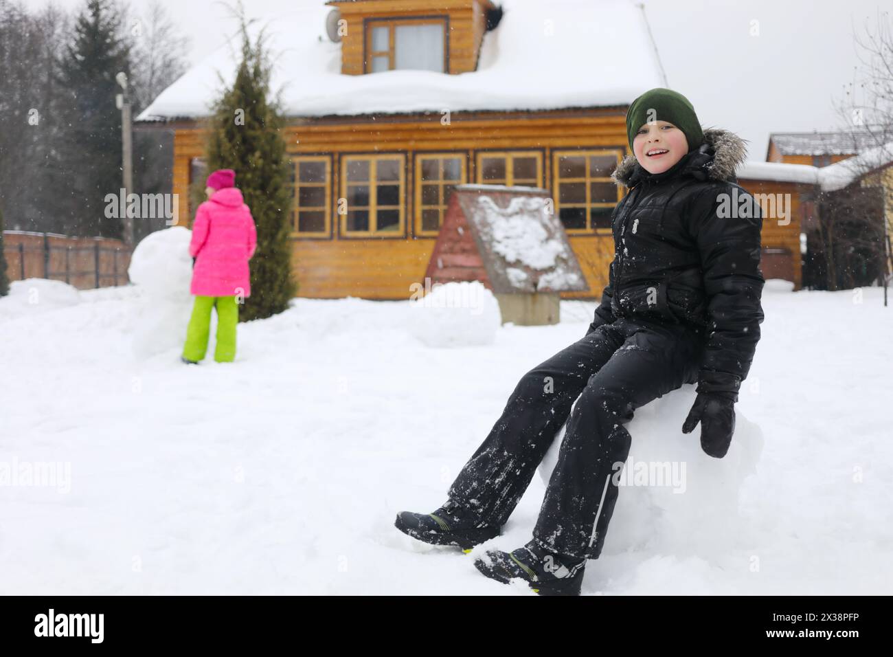 Junge sitzt in einem großen Schneeball, Mädchen macht Schneemänner in der Nähe von hölzernen Landhäusern während des Schneefalls am Wintertag, Mädchen außer Fokus Stockfoto