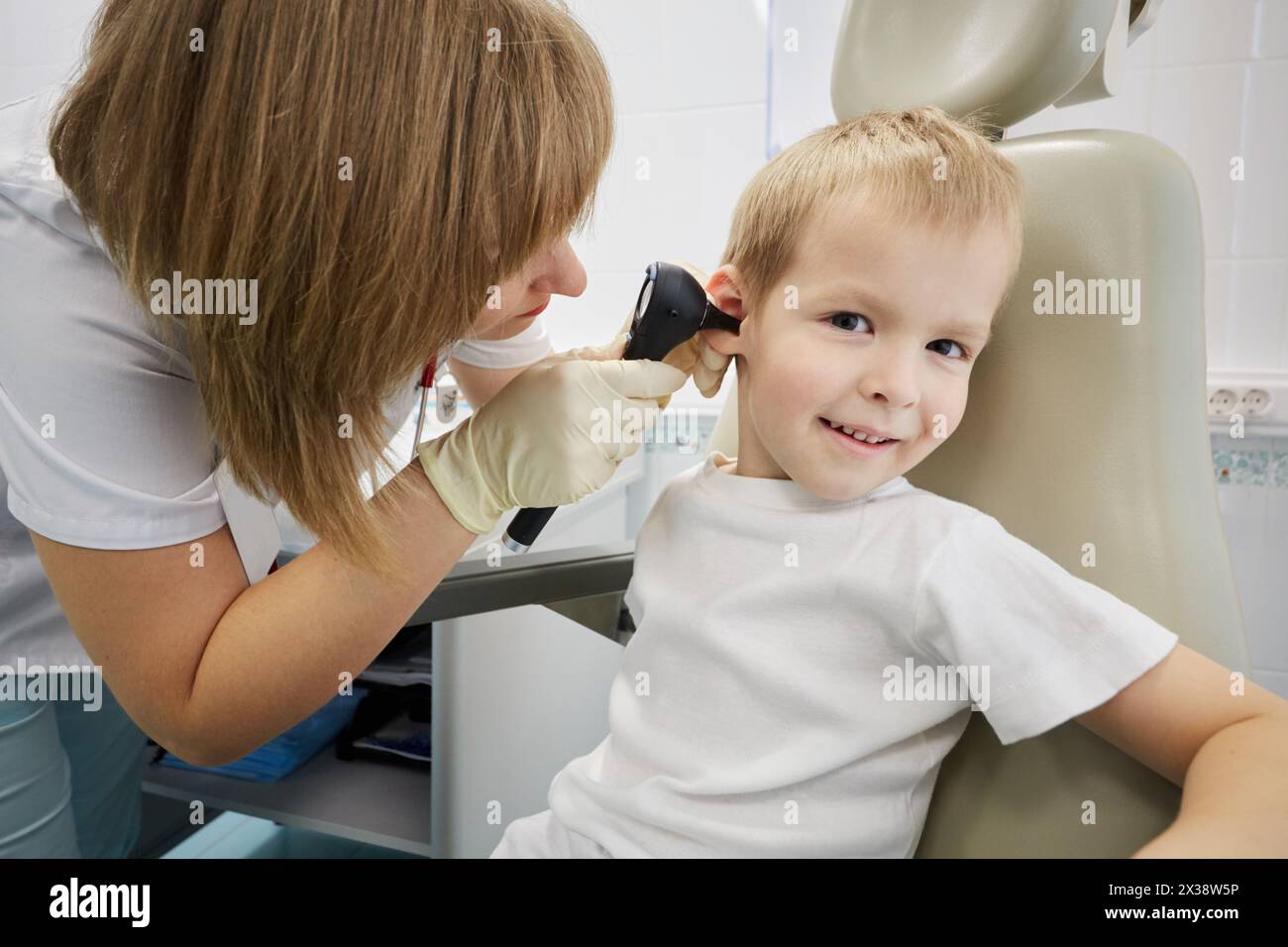 Weibliche Ärztin untersucht das Ohr des lächelnden Jungen, der im medizinischen Sessel sitzt. Stockfoto