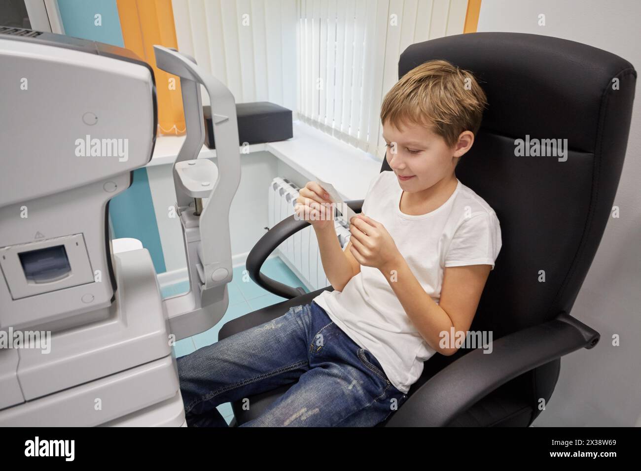 Junge liest Ergebnisse auf Papier, sitzt im Sessel neben Keratometer. Stockfoto
