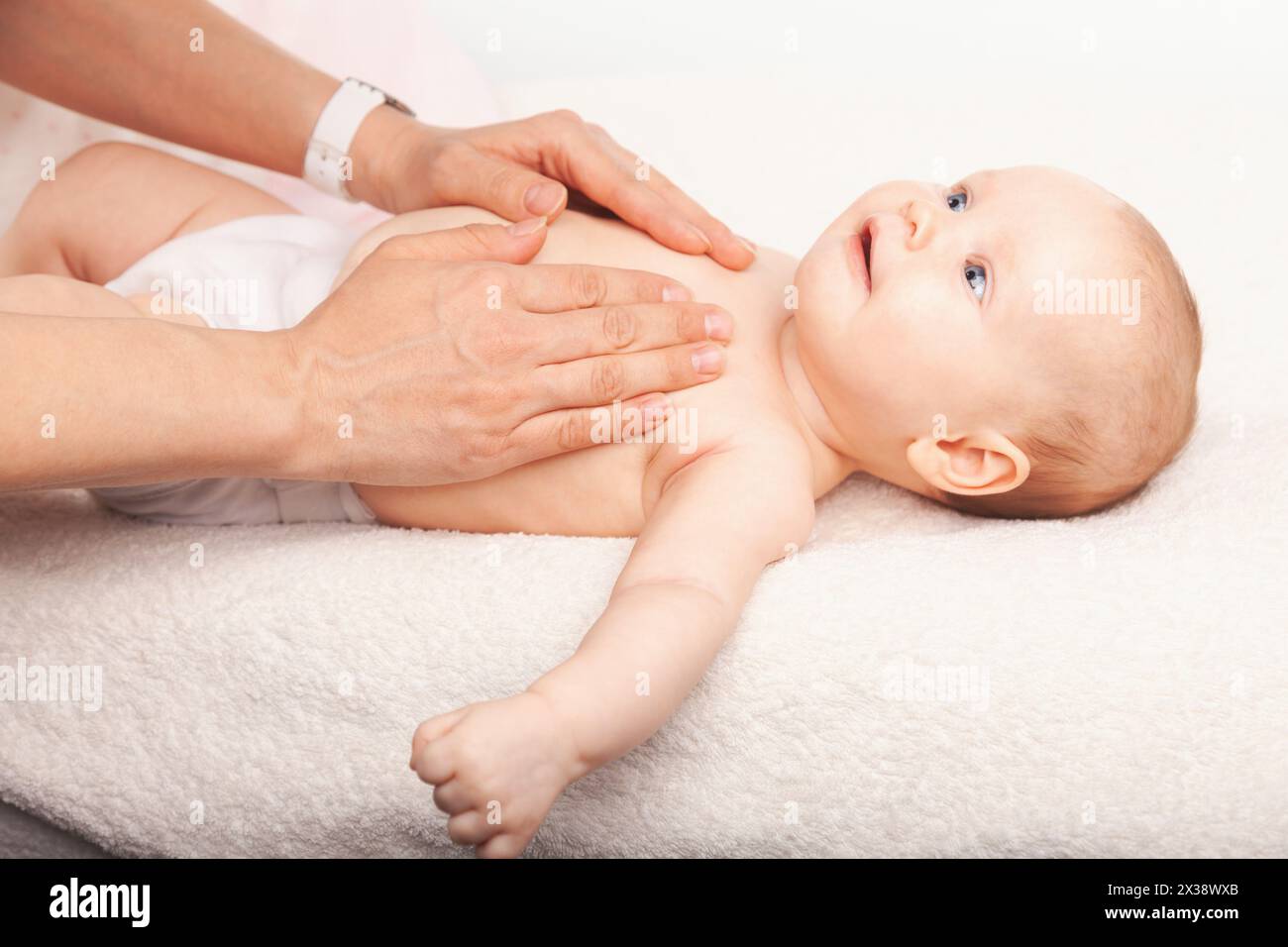 Die Hände eines Pflegepersonals werden sorgfältig gezeigt, um einem zufriedenen Baby, das auf einer weichen weißen Decke liegt, eine wohltuende Brustmassage zu ermöglichen. Die hellen Augen des Säuglings Stockfoto