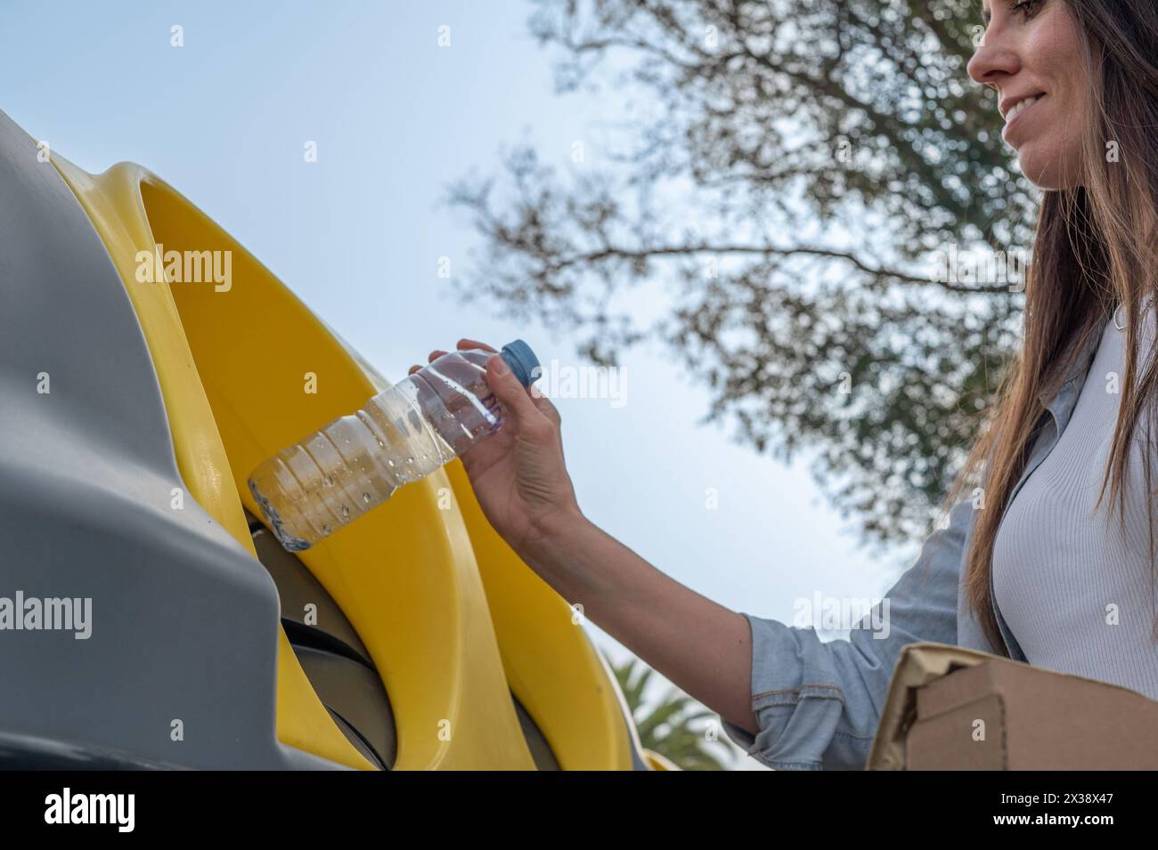 Eine Frau wirft eine Plastikflasche in einen Recyclingbehälter. Die Flasche besteht aus Kunststoff und wird recycelt. Die Frau lächelt beim Werfen Stockfoto