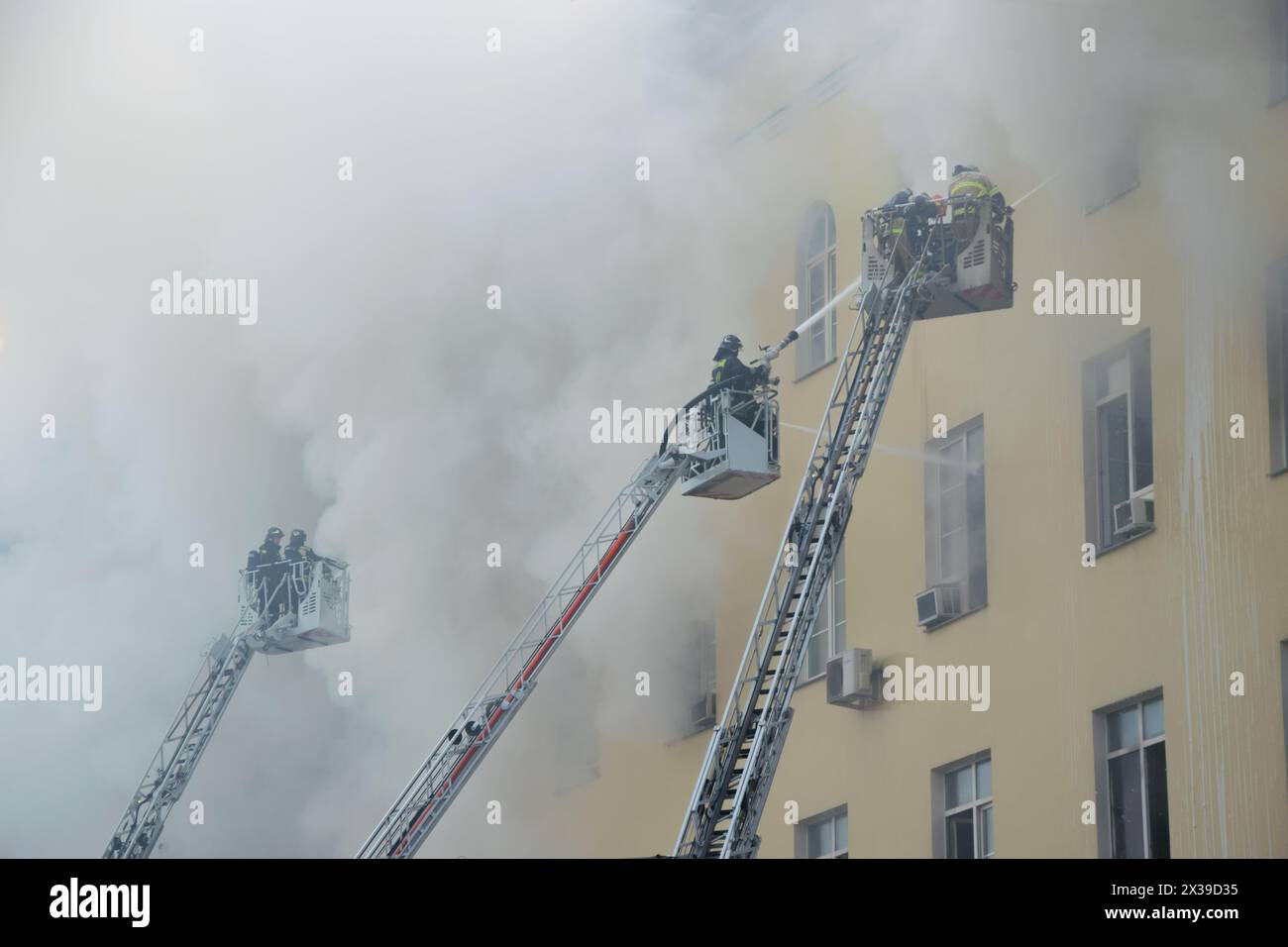 Feuerwehrleute löschen Feuer auf den Leitern von Löschfahrzeugen – gelbes Gebäude in Rauch Stockfoto