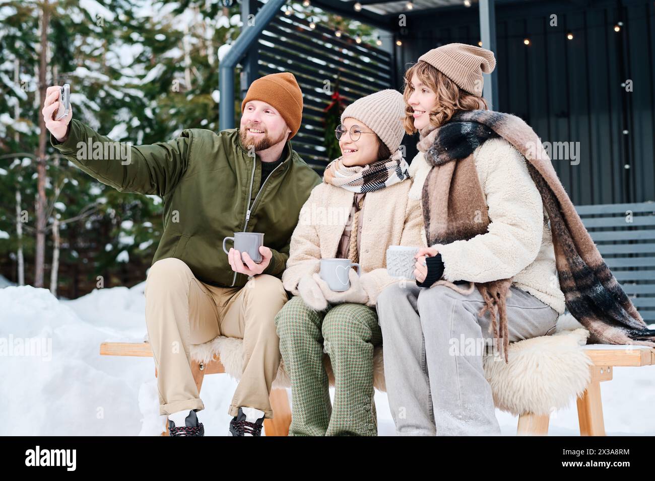 Mittellange Aufnahme einer glücklichen Familie mit Kind, die Fotos auf dem Smartphone macht, während sie am Wintertag im Freien auf der Bank sitzt Stockfoto