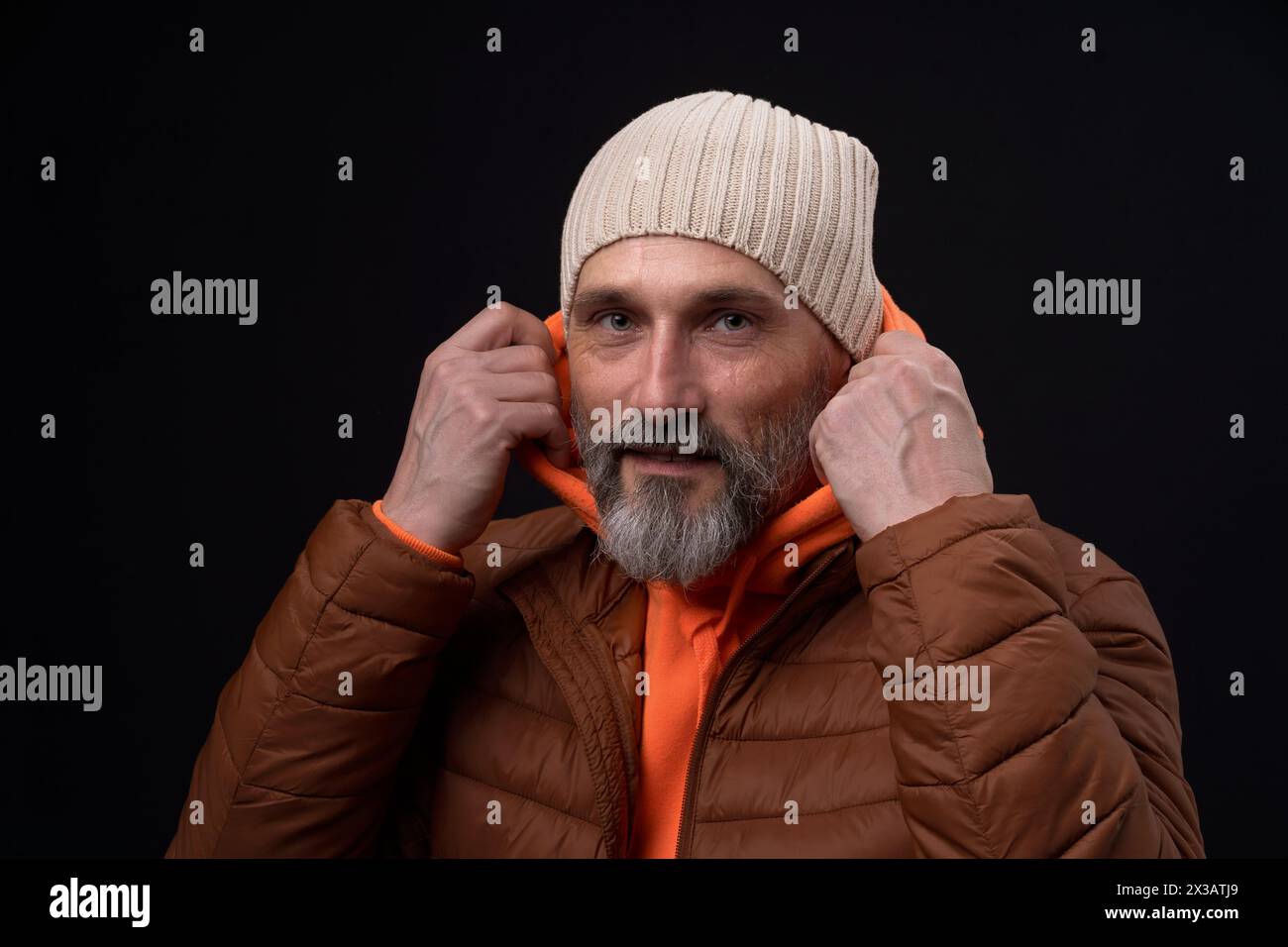 Ein Mann in brauner Jacke und orangefarbenem Hoodie lächelt und passt seinen Hut an. Das Konzept von Wärme und Komfort, da der Mann für kaltes Wetter gekleidet ist Stockfoto