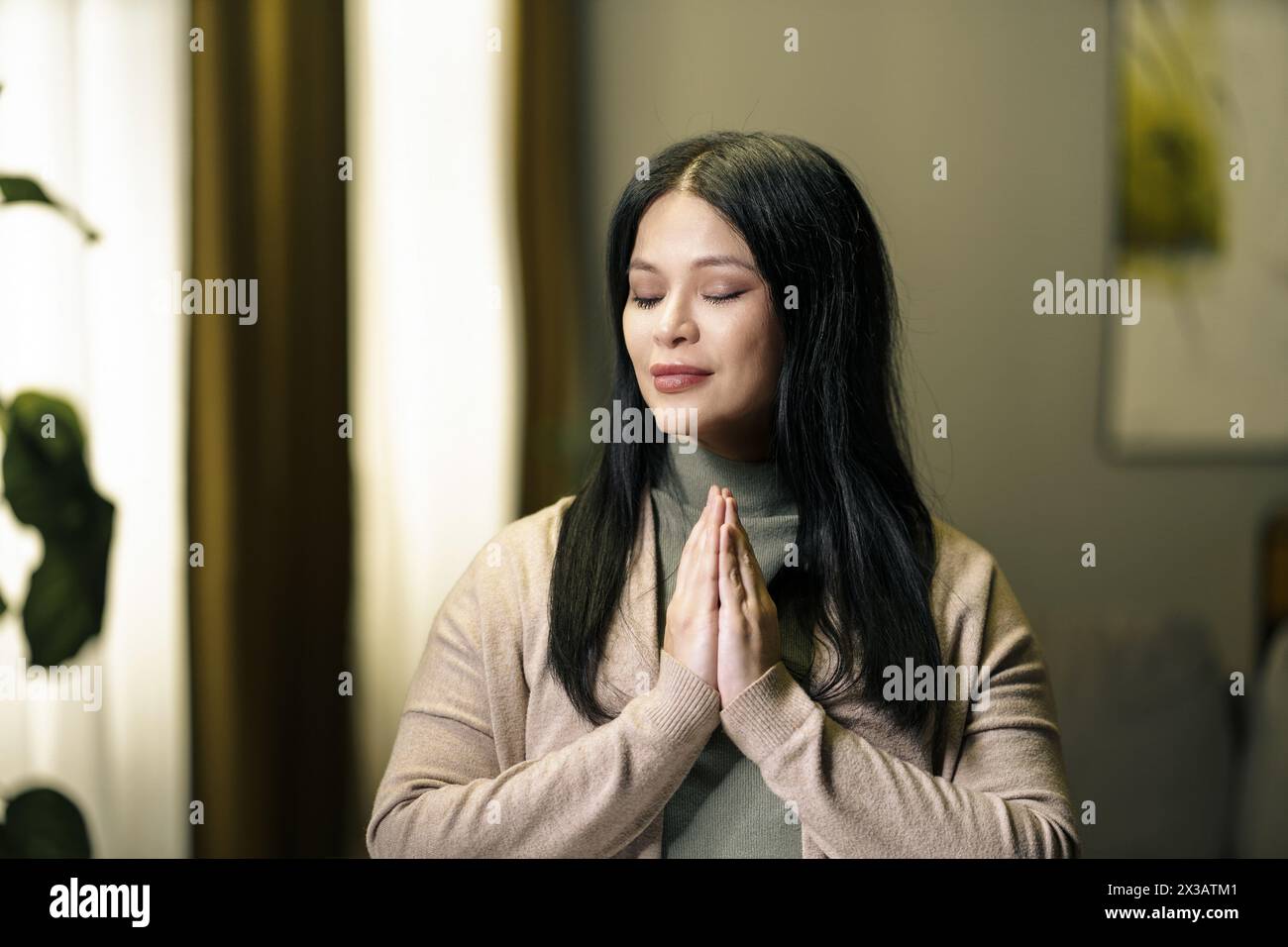 Eine Frau betet in einem Raum mit einer Pflanze im Hintergrund. Sie trägt einen braunen Pullover und hat ihre Hände zusammengelegt Stockfoto