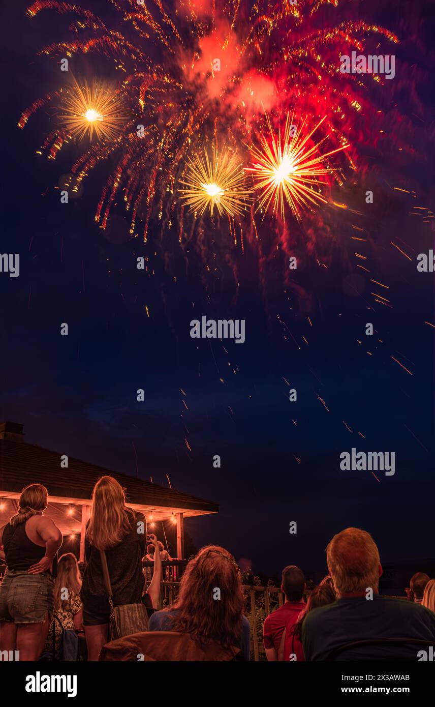 Feuerwerk am 4. Juli in einer kleinen Stadt im Mittleren Westen; Figuren von Menschen, die im Vordergrund sitzen und stehen; beleuchteter Pavillon im Hintergrund Stockfoto
