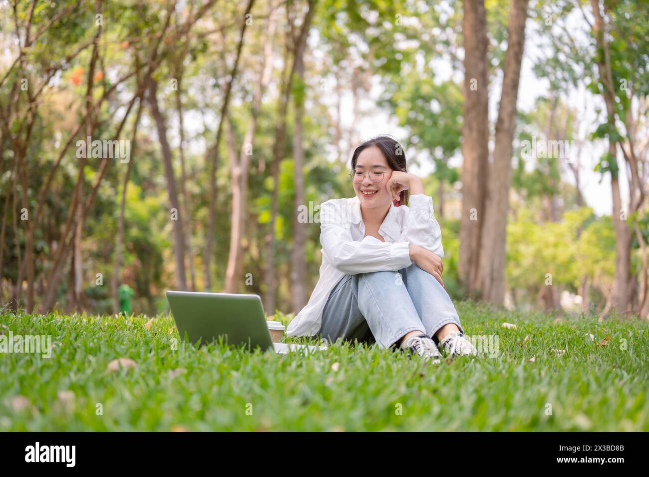 Eine fröhliche junge Frau mit Brille sitzt auf dem Gras in einem Park, lächelt warm und genießt einen Moment der Entspannung mit ihrem Laptop in der Nähe. Wireless-Technologie Stockfoto