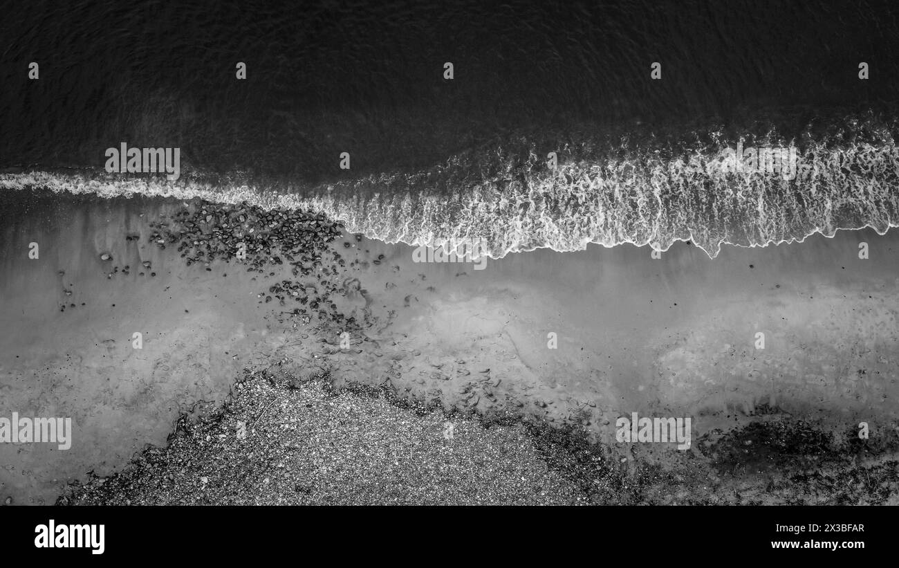 Aus der Vogelperspektive auf eine ruhige Küste mit Wellen, Schaum und Kieselsteinen in Schwarz-weiß, aus der Vogelperspektive auf den Strand Stockfoto