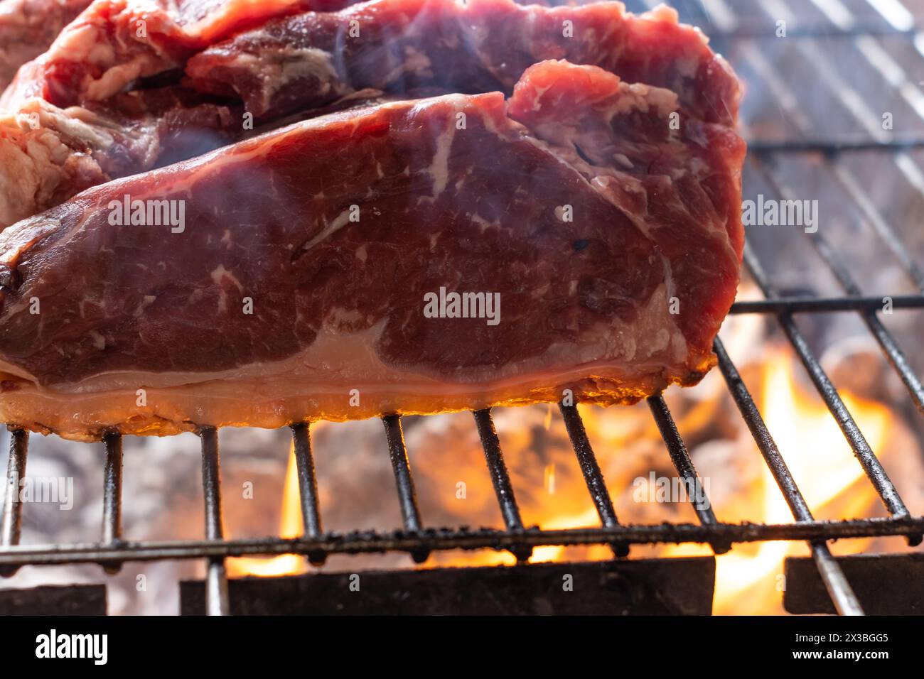 Nahaufnahme von rohen Fleischstücken, die wie Steaks auf einem Grill geschnitten sind, unter dem das Feuer brennt Stockfoto