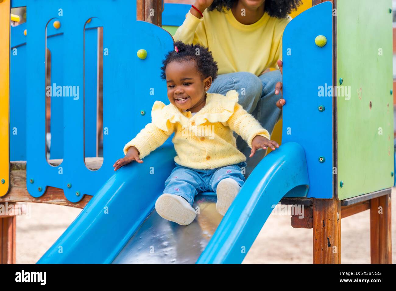 Niedliches afrikanisches Mädchen, das auf einem farbenfrohen Spielplatz neben ihrer Mutter rutscht Stockfoto