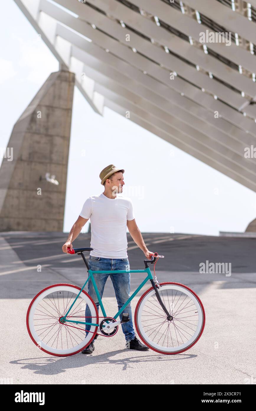 Lässig gekleideter Mann steht mit einem Fahrrad mit fester Ausrüstung und blickt weg, moderne Architektur schafft eine geometrische Kulisse, die an urbane Entdeckungen und styl erinnert Stockfoto