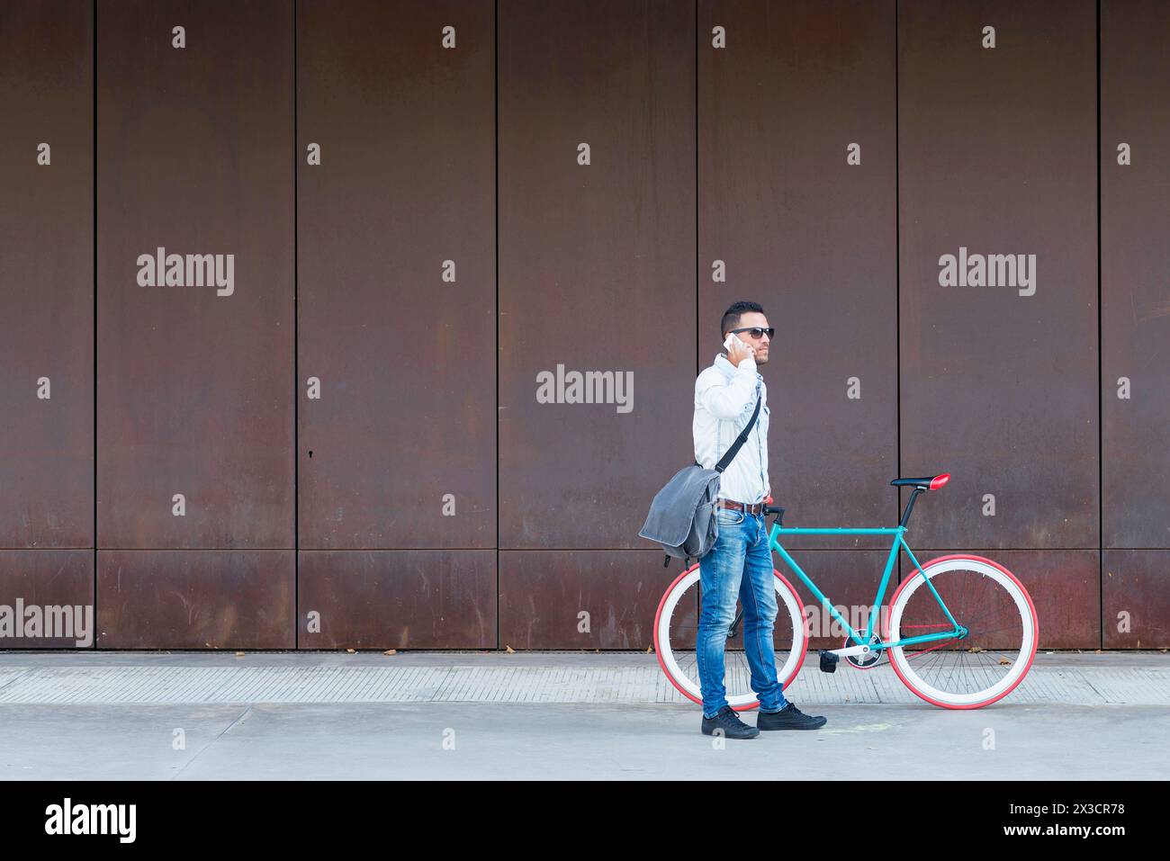 Stilvoller Mann mit Sonnenbrille und Kuriertasche, der neben einem roten Fahrrad steht und einen Anruf gegen eine rostfreie Metallwand führt Stockfoto