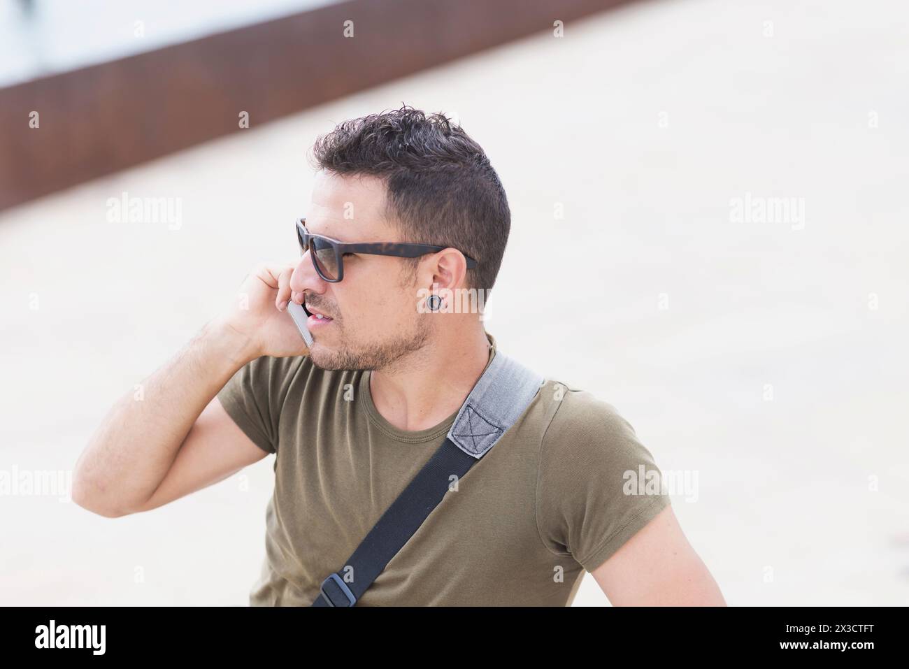 Stilvoller Mann in Sonnenbrille und Kuriertasche, der telefoniert und sich in urbaner Umgebung auf ein Oldtimer-Fahrrad stützt, was auf Mobilität und Stadtleben hindeutet Stockfoto