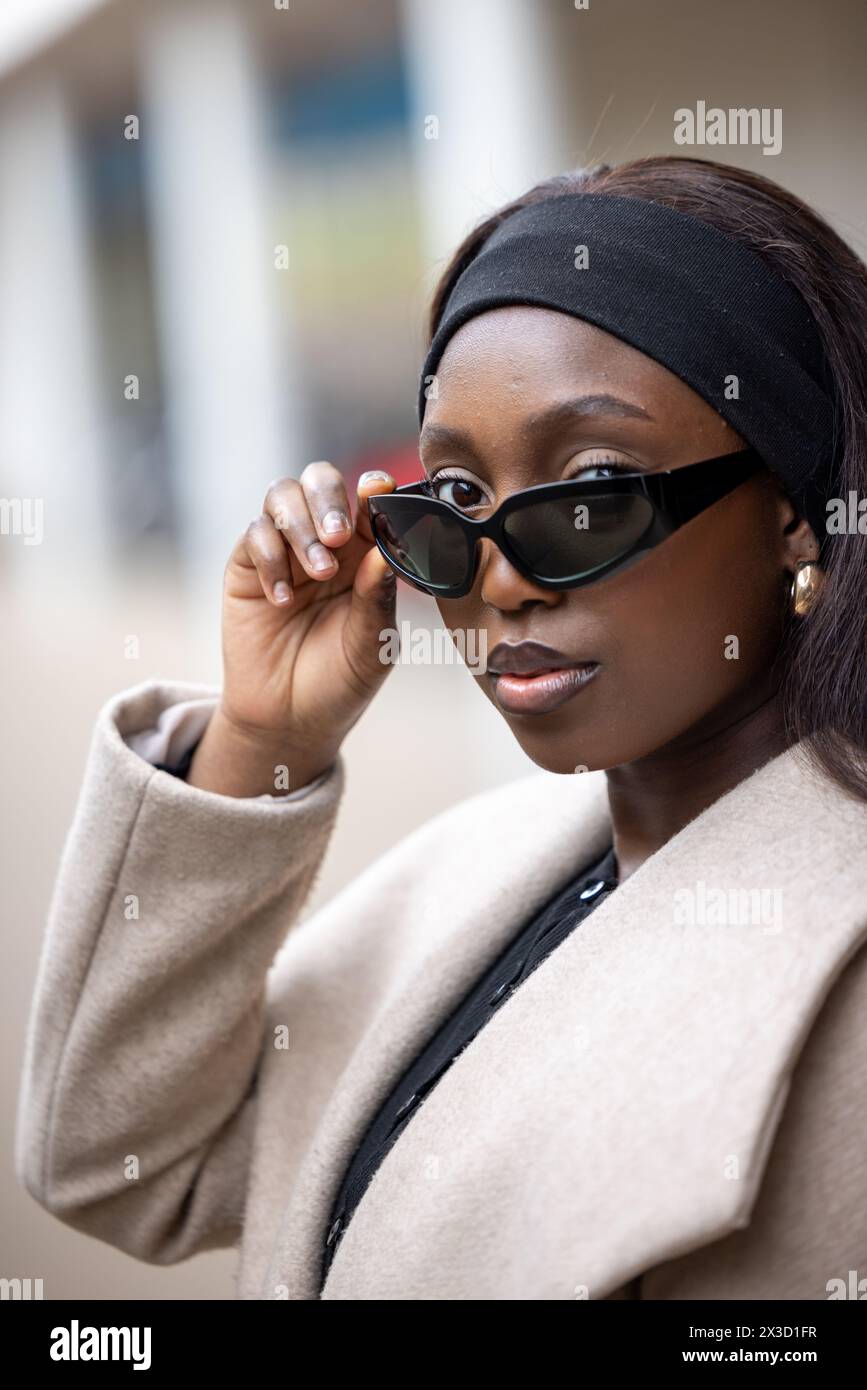 Eine modebewusste schwarze Frau steht auf einer Straße der Stadt, die in dem Moment festgehalten wird, in dem sie ihre elegante Sonnenbrille einstellt. Ihr beigefarbener Mantel und ihr Stirnband verkörpern urbanen Chic, während ihr bewusstes Auftreten eine Mischung aus Raffinesse und Zugänglichkeit widerspiegelt. Das verschwommene Stadtbild im Hintergrund lenkt die Aufmerksamkeit auf ihr stilvolles Ensemble und das coole Selbstvertrauen in ihrem Ausdruck. Stilvolle Frau in Beige Sonnenbrille mit verstellbarem Mantel auf der City Street. Hochwertige Fotos Stockfoto