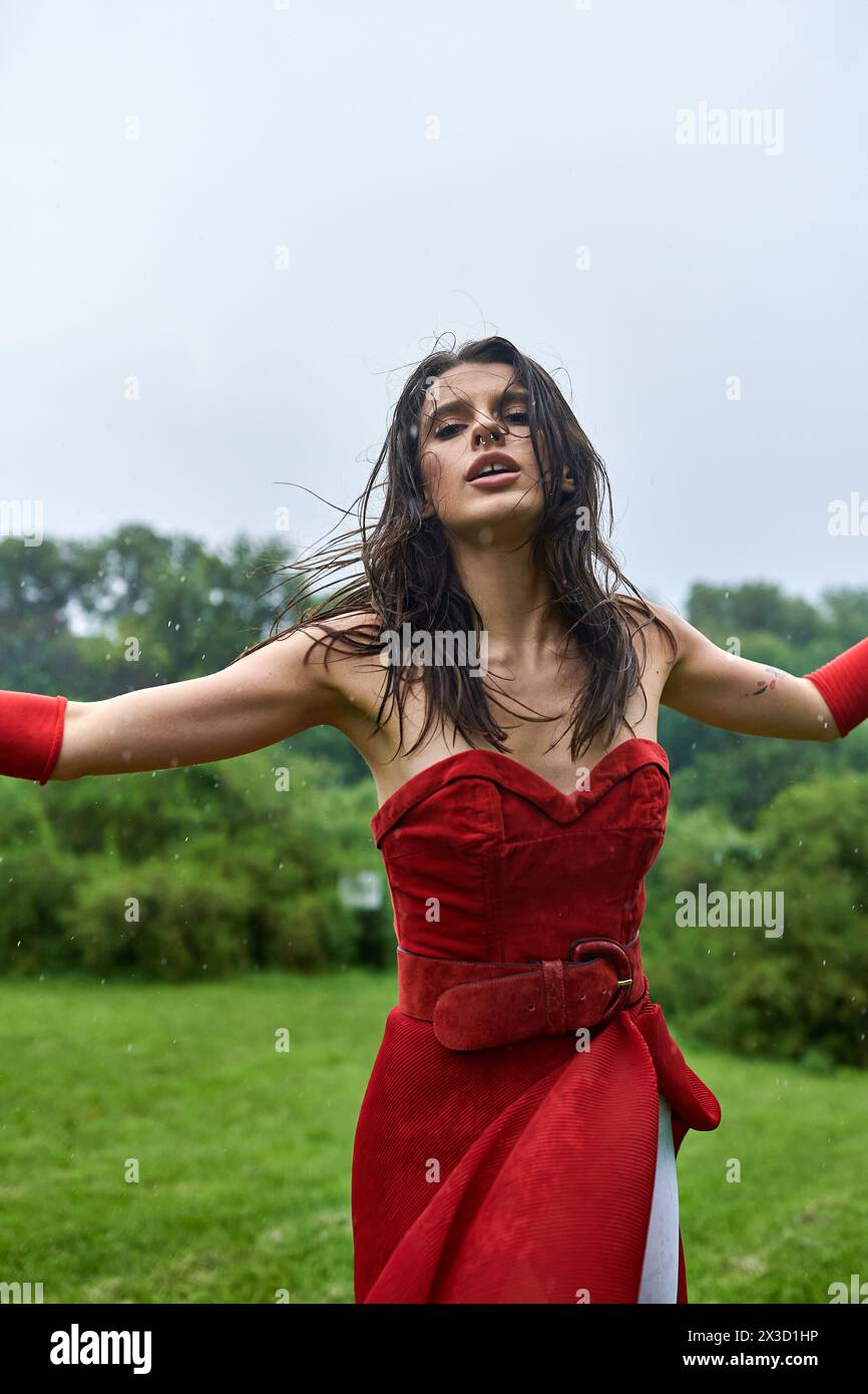 Eine atemberaubende junge Frau in einem roten Kleid und langen Handschuhen steht anmutig auf einem Feld und spürt die sanfte Sommerbrise. Stockfoto
