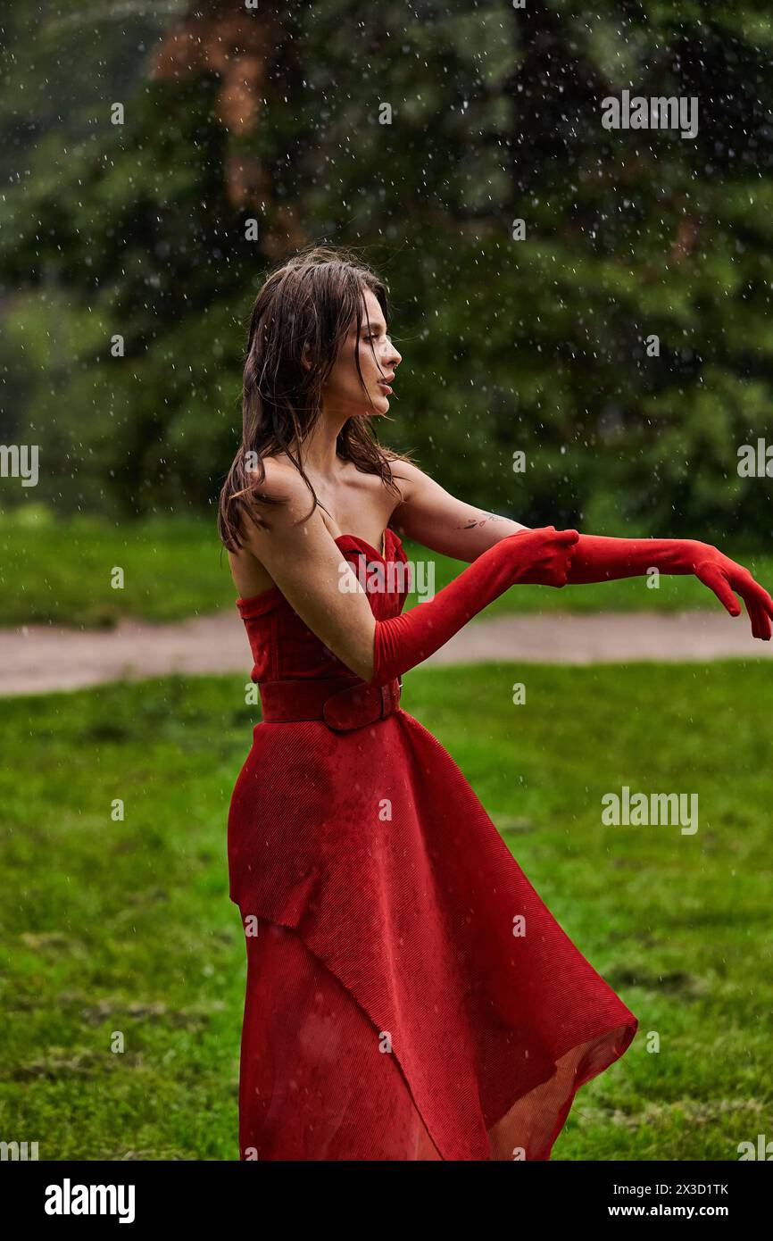 Eine junge Frau in fließendem rotem Kleid und langen Handschuhen tanzt anmutig im Sommerregen und verkörpert Eleganz. Stockfoto