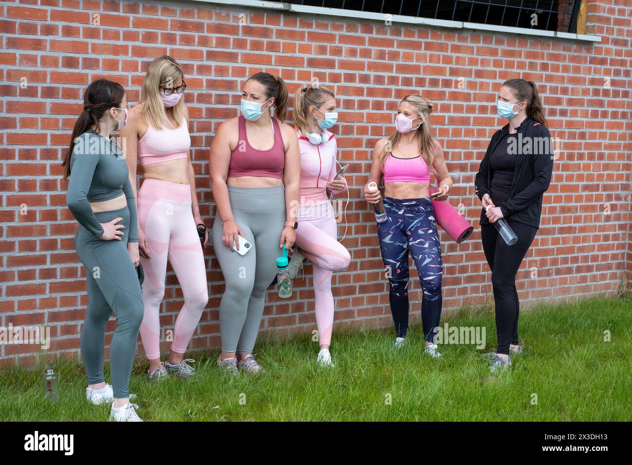 Eine Gruppe von Frauen steht draußen in einer legeren Gemütlichkeit an einer Ziegelwand und trägt Sportbekleidung und Schutzmasken. Sie scheinen ein Gespräch zu führen und möglicherweise über ihre Fitnessroutine oder die einzigartigen Herausforderungen zu sprechen, die es mit sich bringt, in Zeiten aktiv zu bleiben, in denen Gesichtsbezüge erforderlich sind. Gruppe von Frauen in Sportswear mit Masken über Outdoor von Brick Wall. Hochwertige Fotos Stockfoto