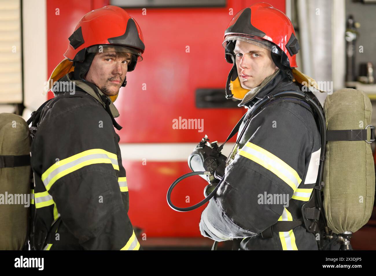 Zwei Feuerwehrmänner in Schutzanzügen und rotem Feuerwehrhelm und Sauerstoffflaschen stehen vor dem Feuerwehrauto, Blick von hinten, umsehen Stockfoto