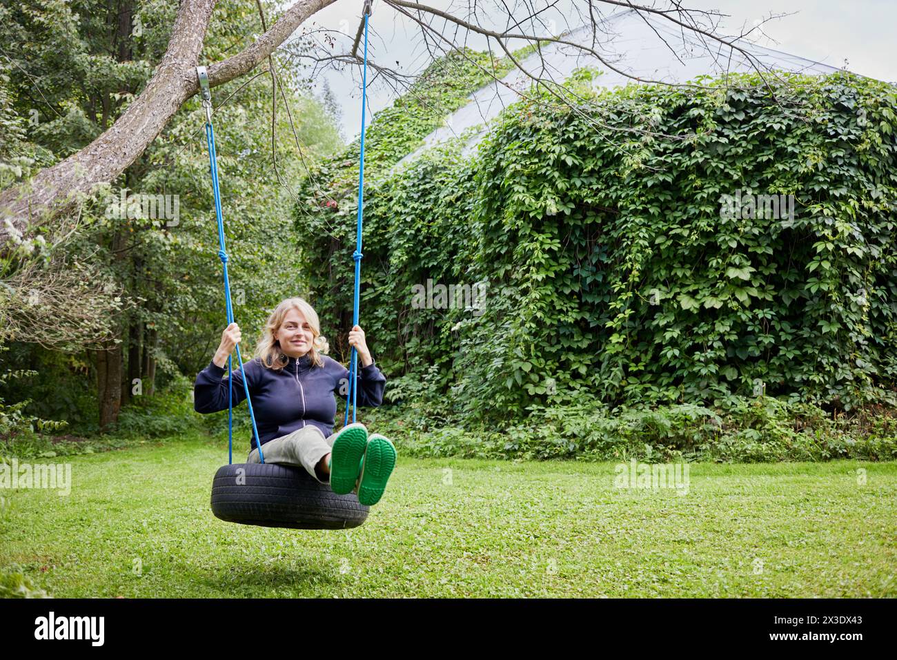 Lächelnde blonde Frau auf Seilschaukeln, die am Baum auf dem Rasen befestigt sind. Stockfoto