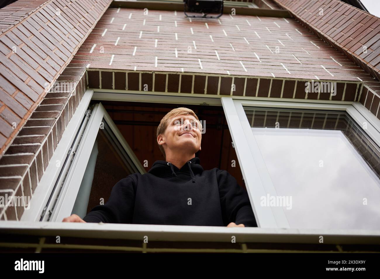 Ein lächelnder junger Mann schaut aus dem Fenster eines Backsteinhauses, aus einem tiefen Winkel. Stockfoto