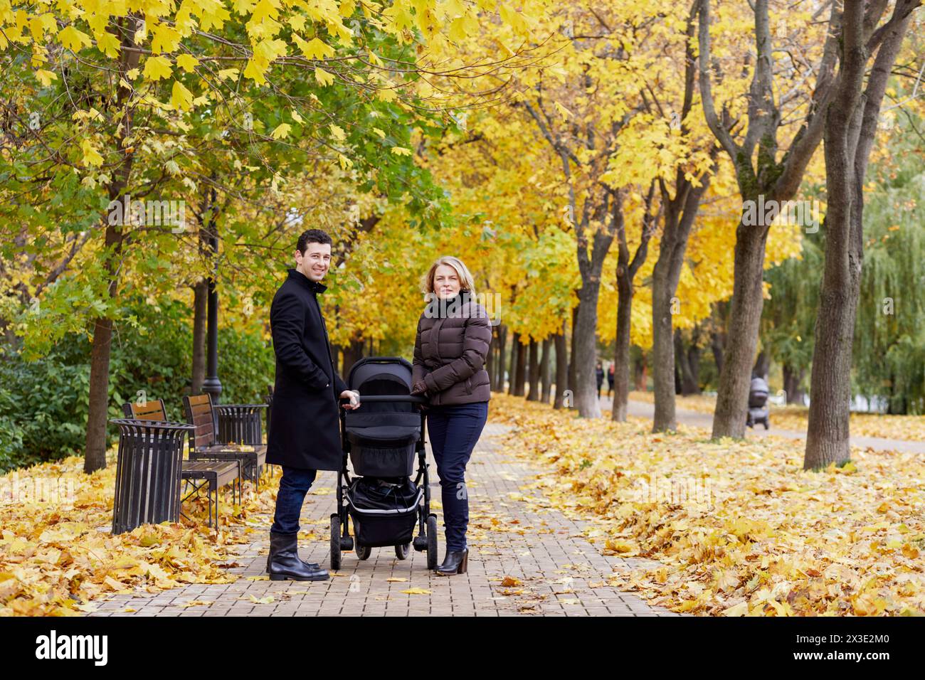 Lächelnder Mann und Frau stehen mit Perambulator im Herbstpark auf dem Weg. Stockfoto