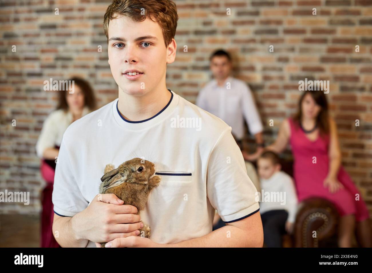 Porträt eines Teenagers, der Kaninchen gegen Familie hält, die auf Sofa im Zimmer sitzt. Stockfoto