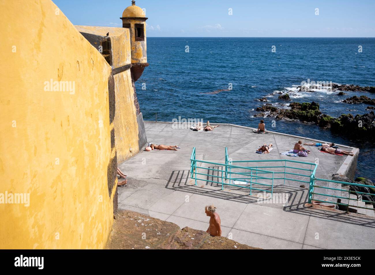 Sonnenanbeter und Schwimmer genießen die Nachmittagssonne auf der Terrasse unter dem Fort Sao Tiago (Gelb) am 19. April 2024 in Funchal, Madeira, Portugal. Ursprünglich erbaut, um die Küste vor Piraterie zu schützen, während des 16. Bis 17. Jahrhunderts unter Philipp I. von Portugal. Das Fort wurde für viele Zwecke genutzt, einschließlich der Unterbringung britischer Truppen während des Halbinsel-Krieges, der Militärpolizei und der Funchal Lancers Squad zu verschiedenen Zeiten. Stockfoto