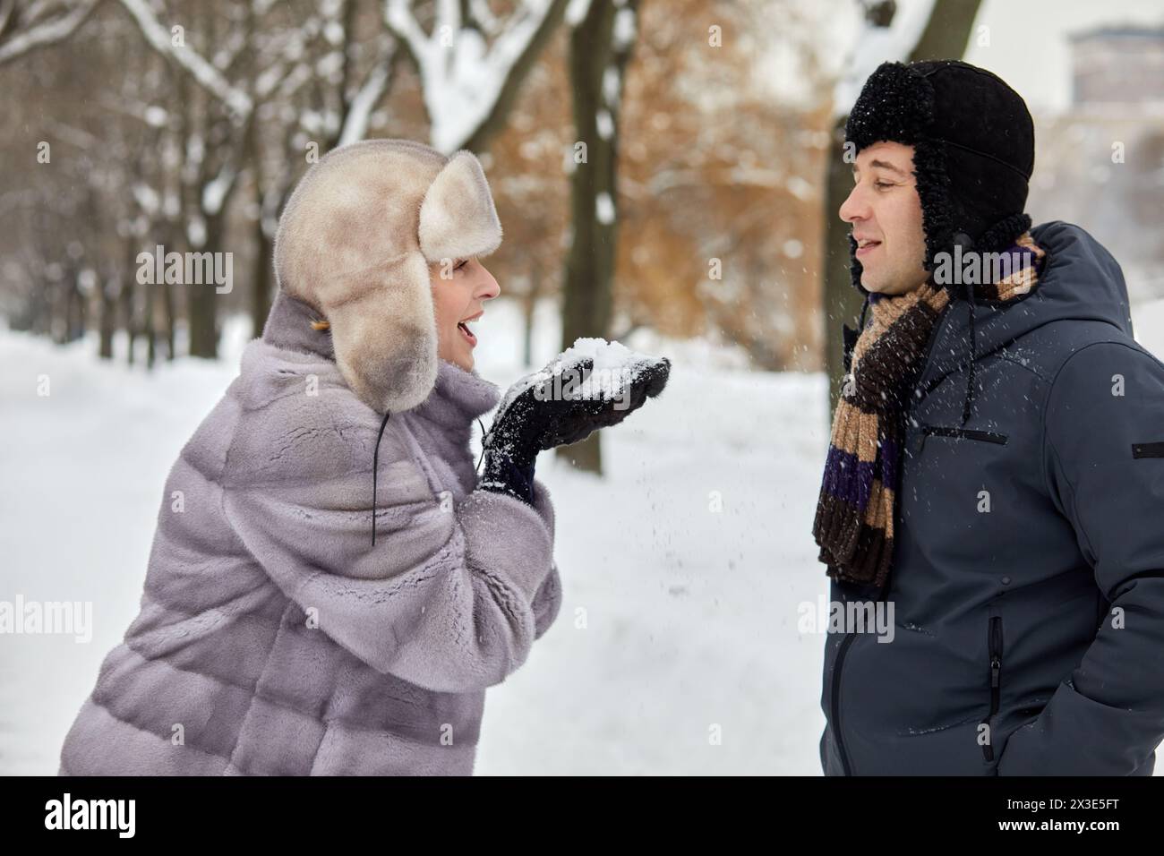 Lächelnde Frau in Pelzmantel und Hut, die Schnee von Fäustlingen auf den Mann gegenüber im Winterpark pustet. Stockfoto