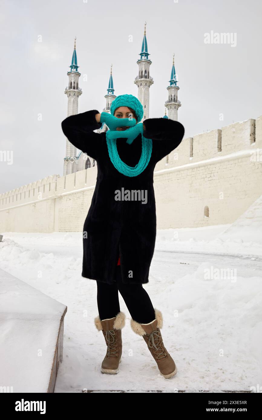 Frau in schwarzem Pelzmantel steht am Wintertag hinter der Wand, die Palmen in gestrickten Fäustlingen in der Nähe des Gesichts gegen die Moschee von Kasan Kul Sharif faltet. Stockfoto