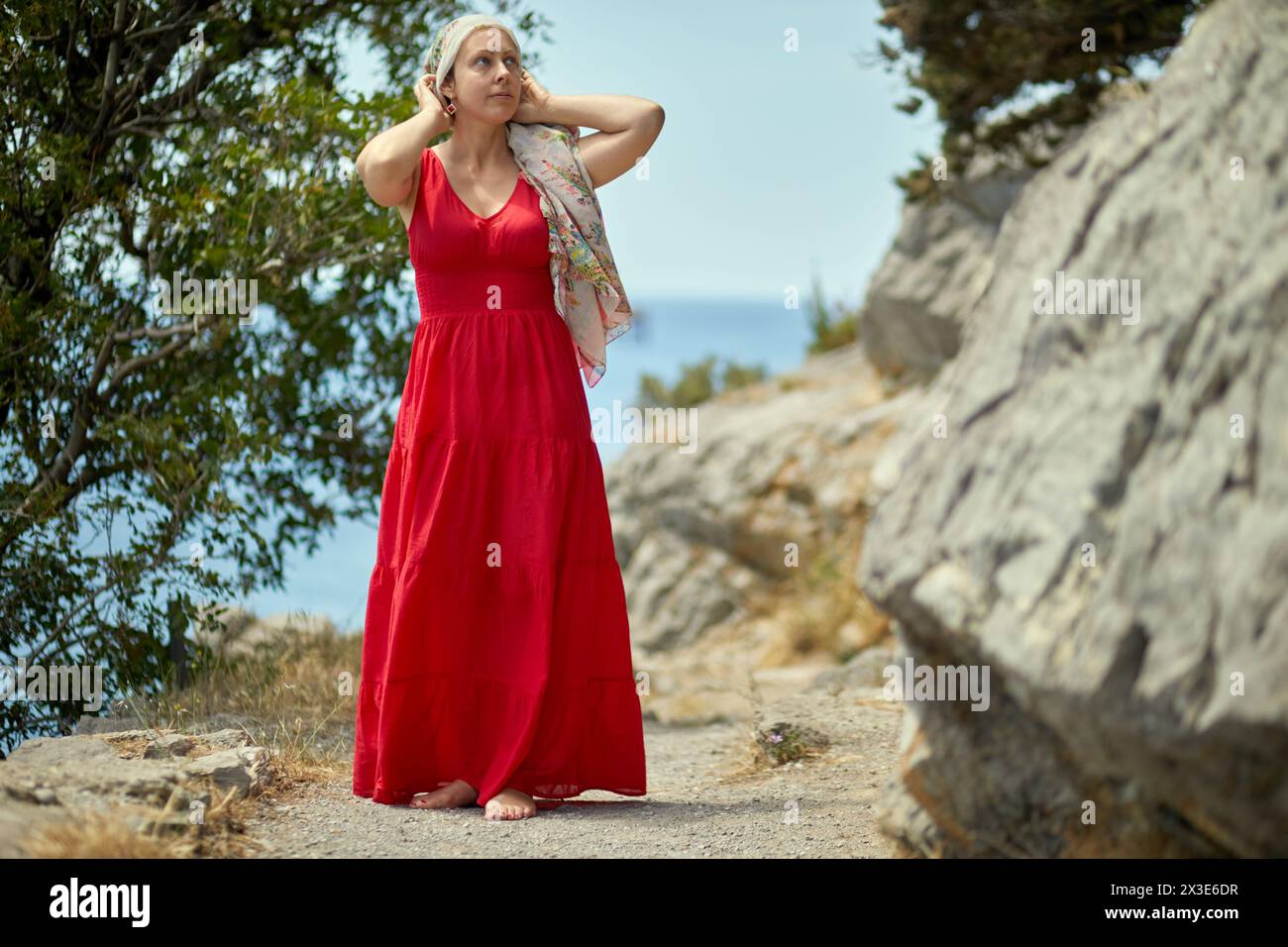 Frau in rotem Kleid und blumigem Schal steht auf einer Klippe über dem Meer. Stockfoto