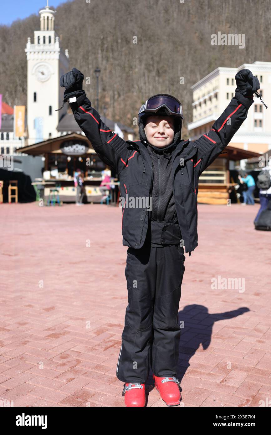 Junge in Helm und Skianzug steht mit erhobenen Armen und geschlossenen Augen auf Platz im Skigebiet mit Bergen, Krasnaja Polyana, Sotschi, Russland Stockfoto