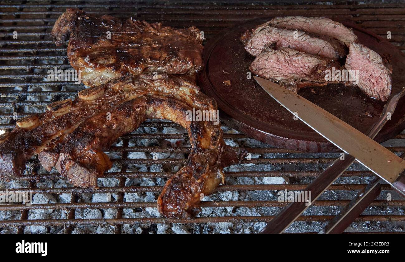 Argentinisches Barbecue. Einige ausgezeichnete Stücke argentinisches Rindfleisch auf einem Holzkohlegrill und einem Holztisch mit Messer. Nahaufnahme der geschnittenen gegrillten Be Stockfoto