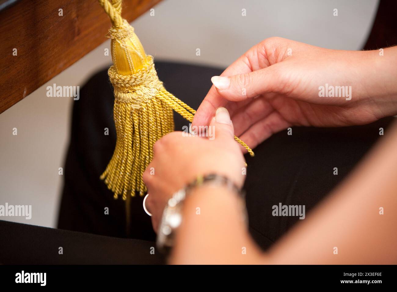 Das Foto bietet einen detaillierten Blick auf die Hände, die eine goldene Quastenschnur akribisch binden, und konzentriert sich dabei auf die Feinheiten der Aufgabe. Die Zeiger sind mit einer Uhr und Schmuck verziert, was eine Vorbereitung auf ein bedeutendes Ereignis andeutet. Die reichhaltige Textur der Quaste verleiht einen luxuriösen Touch. Eleganz beim Basteln: Hände binden ein goldenes Quastenband. Hochwertige Fotos Stockfoto