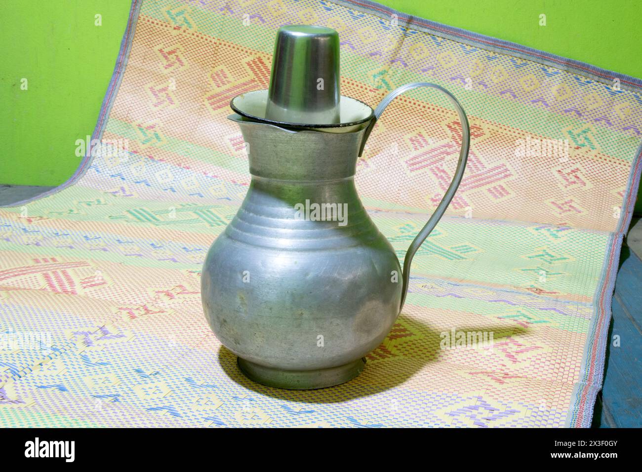 Ein Wasserjacken aus Aluminium mit einem Becher aus Edelstahl oder Glas auf einer Matte. Altmodischer Wassertopf mit Halter oder Handlauf. Häufig in Bangladesch verwendet. Stockfoto