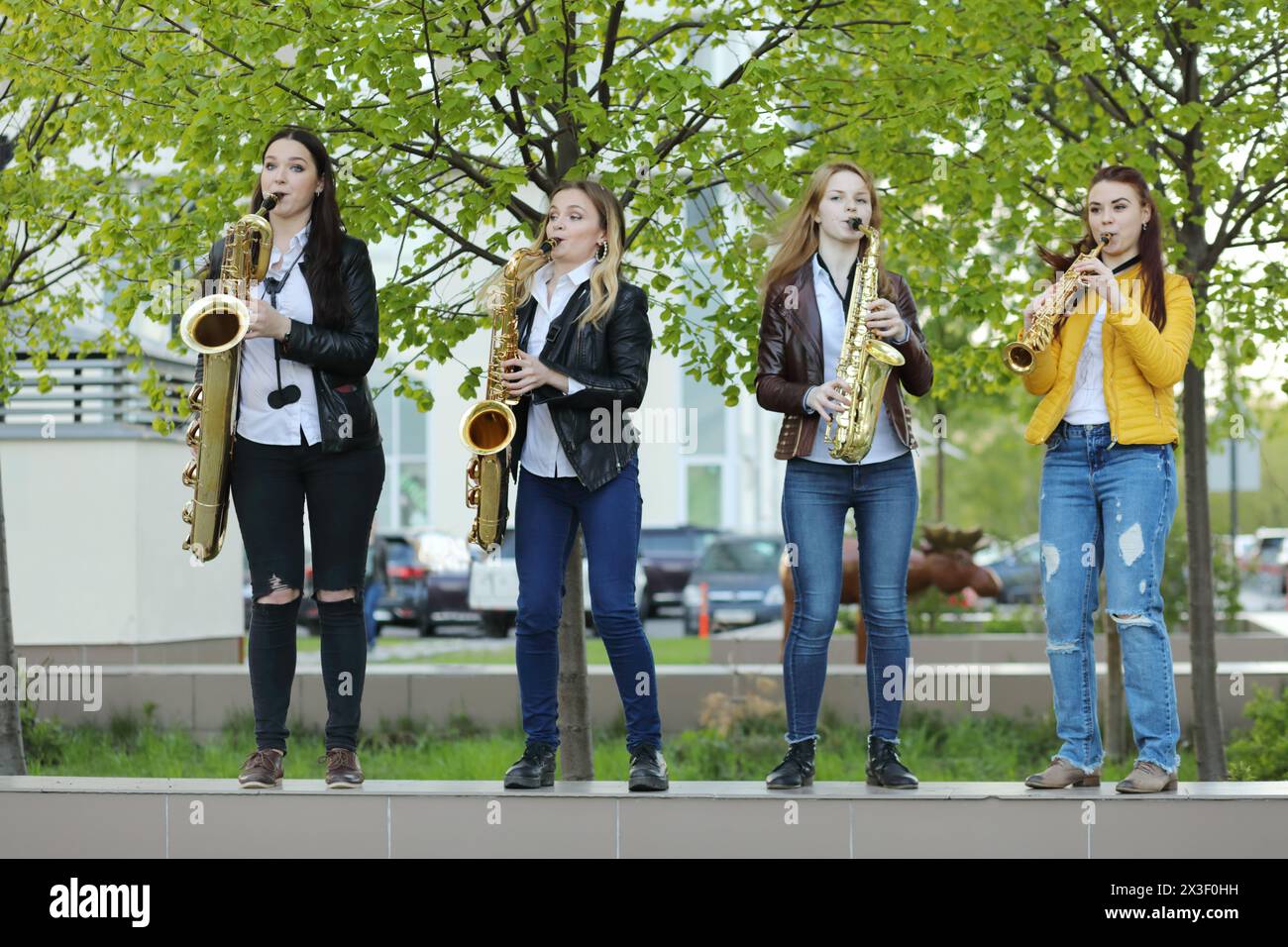 Vier junge hübsche Frauen in Jeans spielen am Frühlingstag im Freien Saxophon Stockfoto