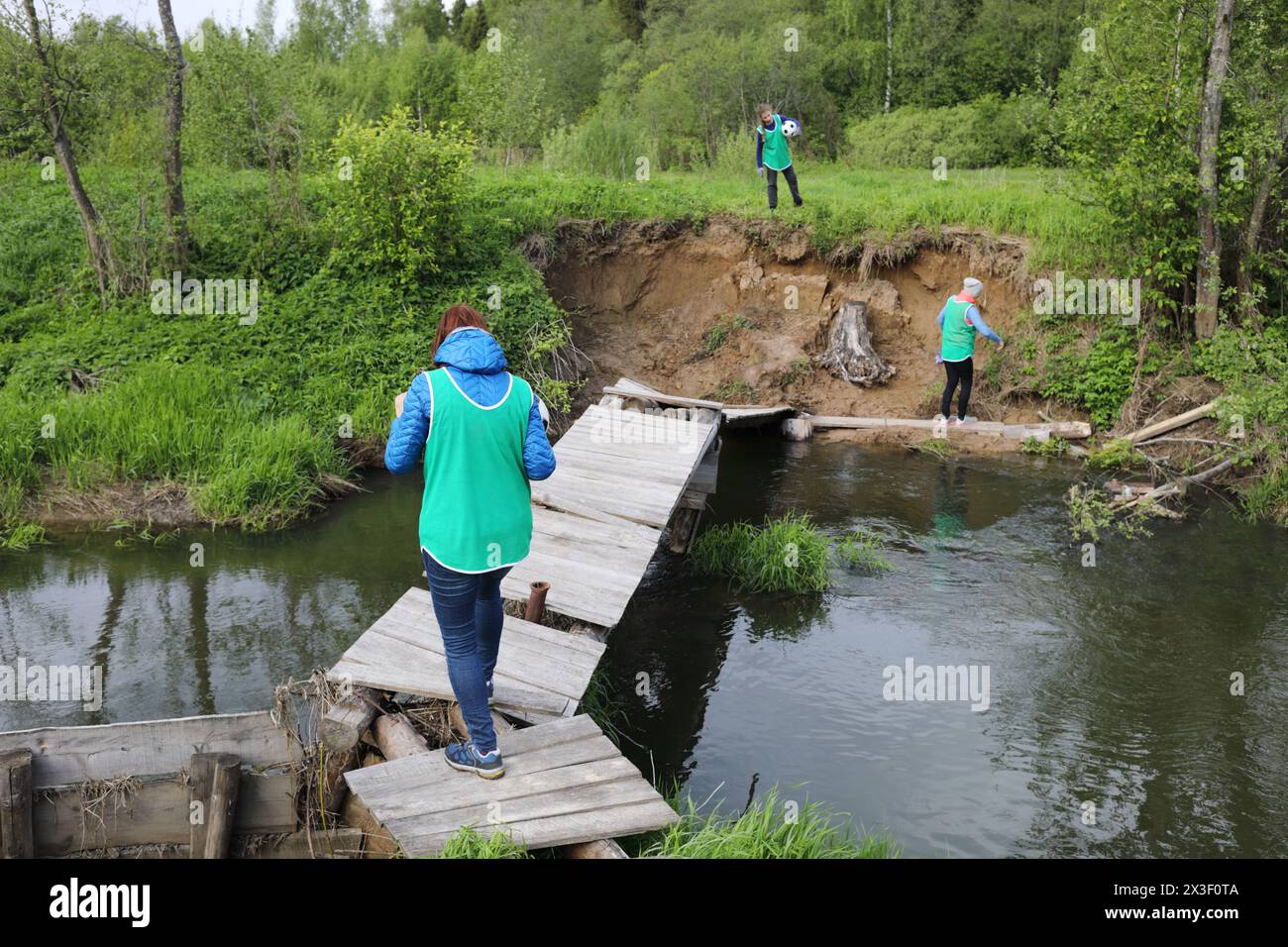 Zwei Frauen und Mädchen gehen auf eine Holzbrücke über einen kleinen Fluss im grünen Wald Stockfoto