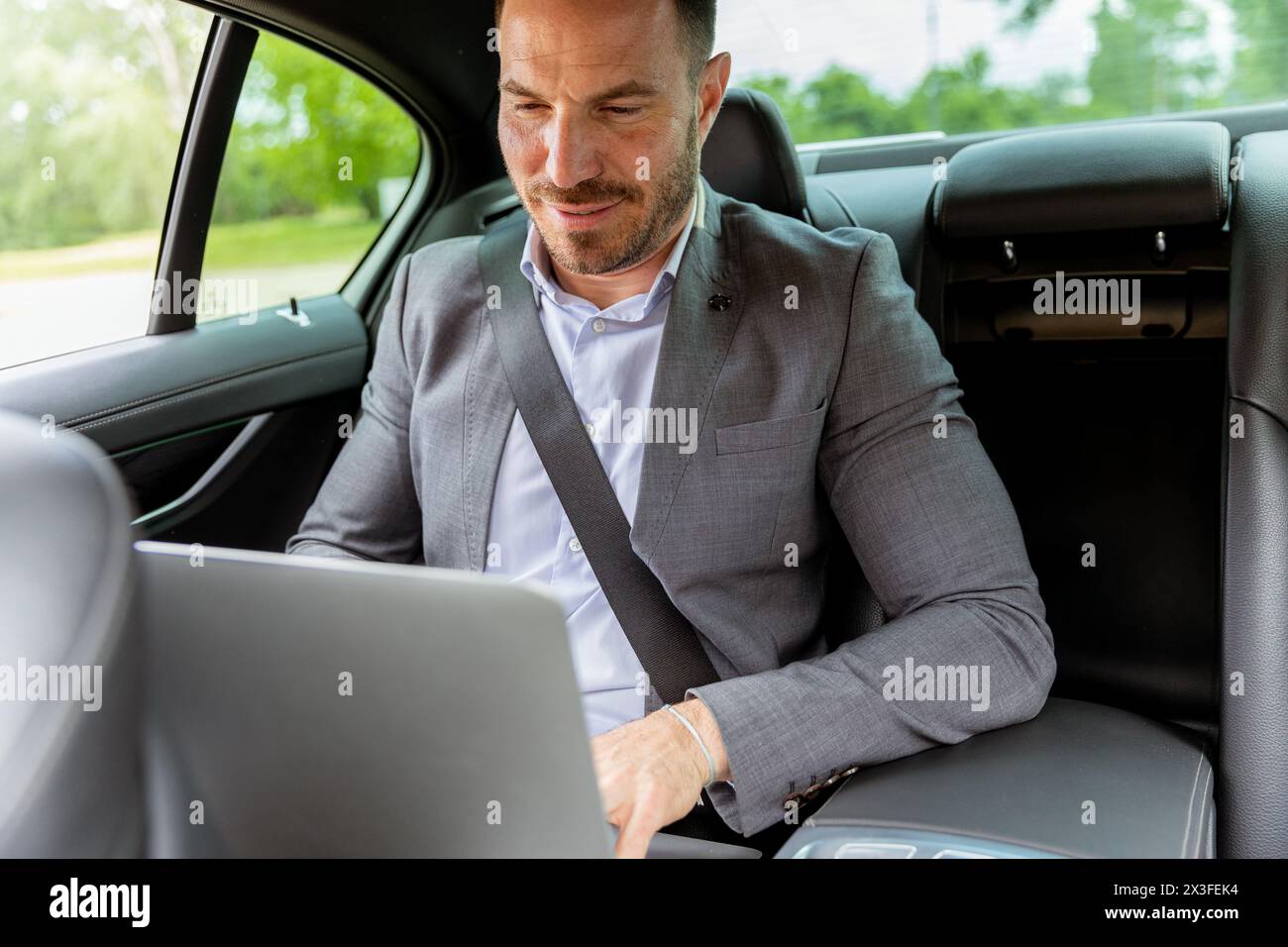 Fokussierter Mann in Geschäftskleidung, der auf einem Laptop auf dem Rücksitz eines Autos schreibt und jede Minute nutzt Stockfoto
