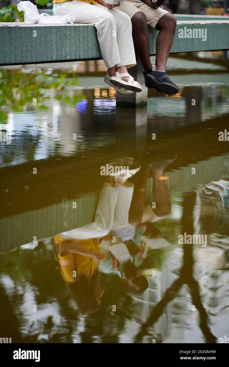 Reflexion über die Wasseroberfläche von jungen Reisenden, die auf architektonischen Bauten in urbaner Umgebung sitzen und eine Pause genießen Stockfoto