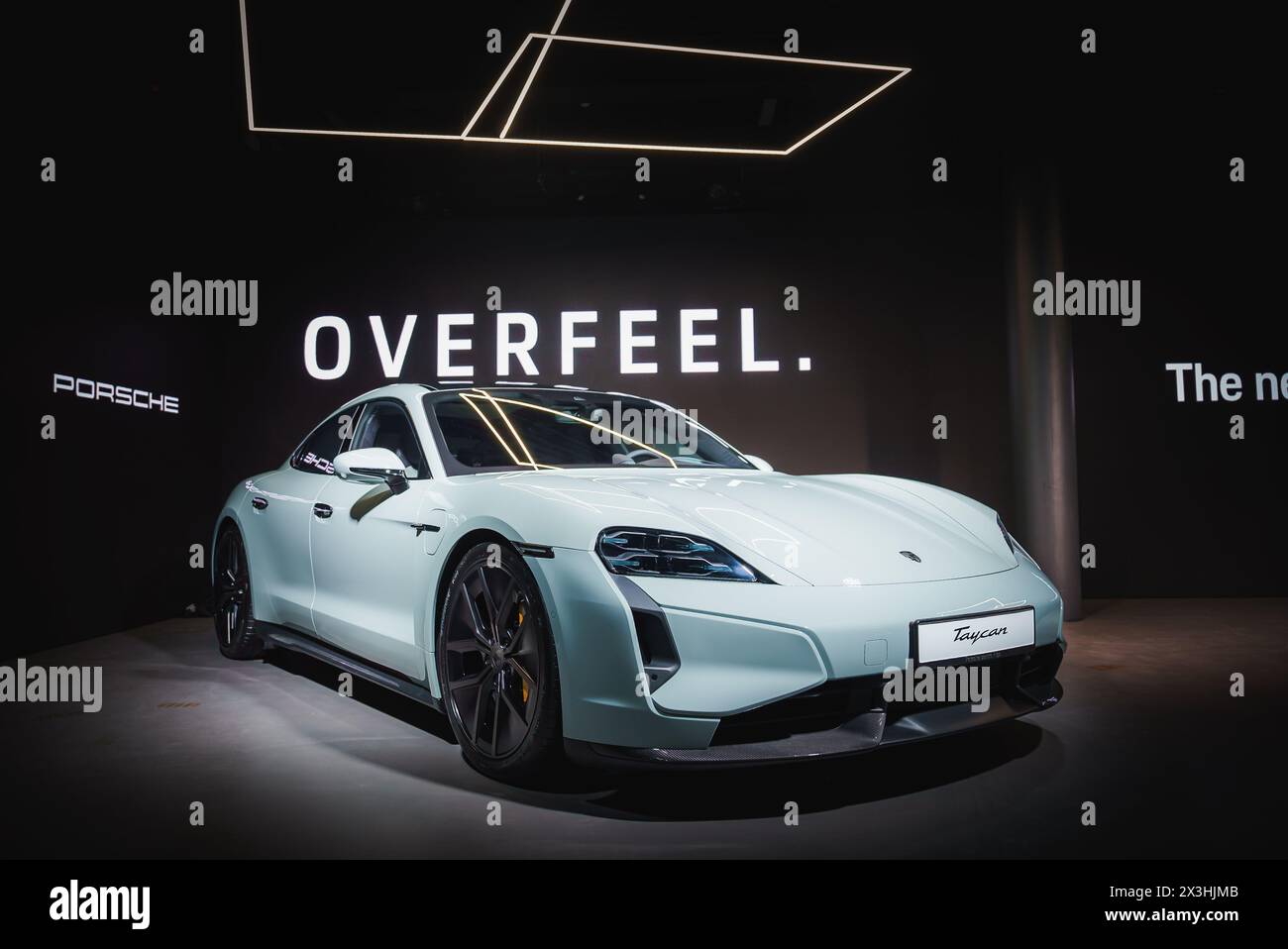 Hellblauer Porsche Taycan auf der Autoshow mit OVERFEEL-Schriftzug im dunklen Raum. Stockfoto