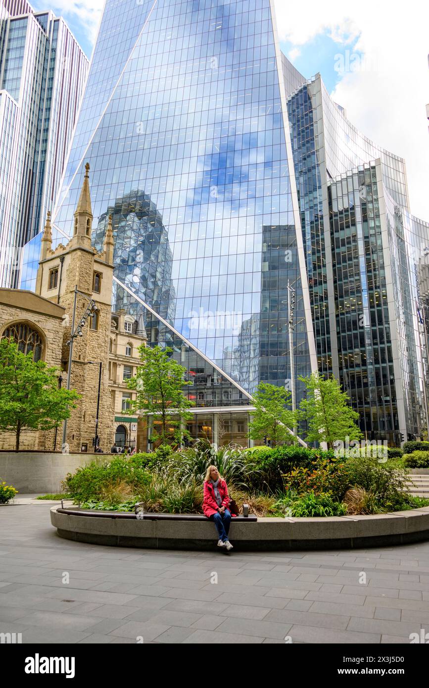 Frau, die in einem grünen Raum zwischen hohen Büroblöcken aus Glas und Stahl in der Nähe der St. Andrew Undershaft Church, Leadenhall Streel, City of London, sitzt Stockfoto