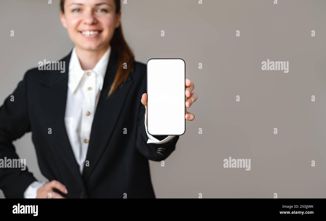 Geschäftsfrau im Anzug zeigt den Bildschirm ihres Smartphones zur Kamera und lächelt. Mockup eines leeren Bildschirms des Mobiltelefons in der Hand des weiblichen Geschäftsführers Stockfoto