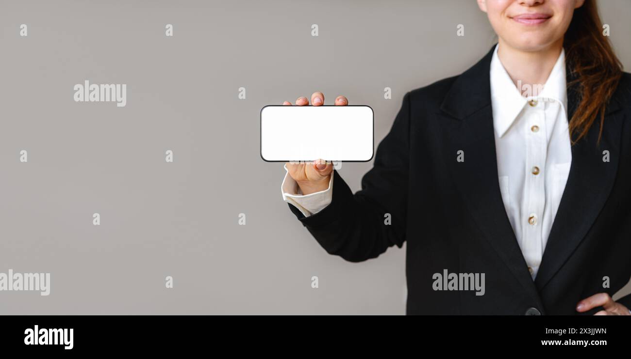 Mockup eines leeren Bildschirms des Mobiltelefons in der Hand einer Geschäftsfrau auf grauem Hintergrund. Geschäftsfrau im Anzug zeigt den Bildschirm ihres Smartphones Stockfoto
