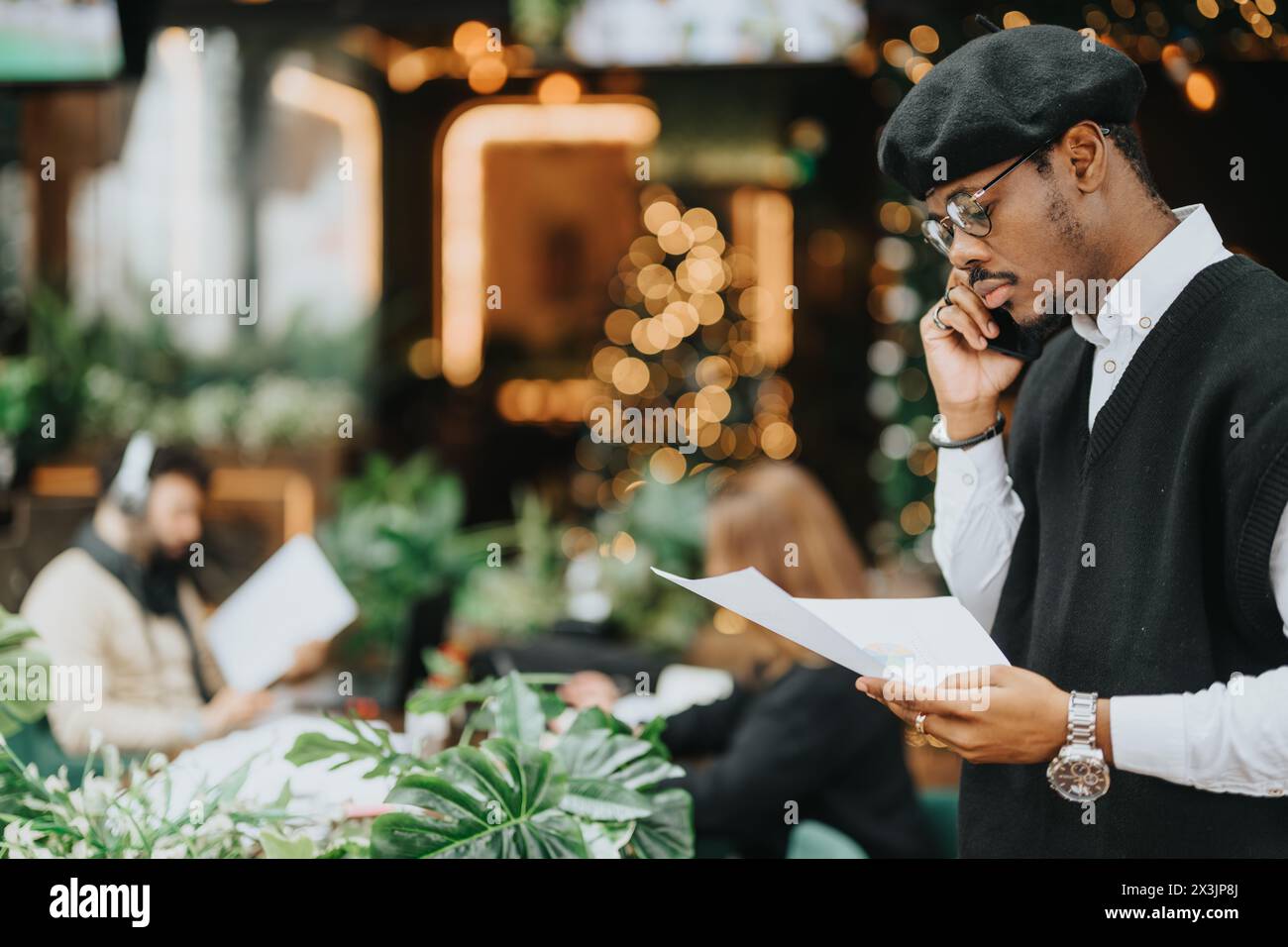 Ein aufmerksamer junger Mann in stilvoller Kleidung bewertet Papierkram, während er in einem üppigen Café telefoniert. Stockfoto