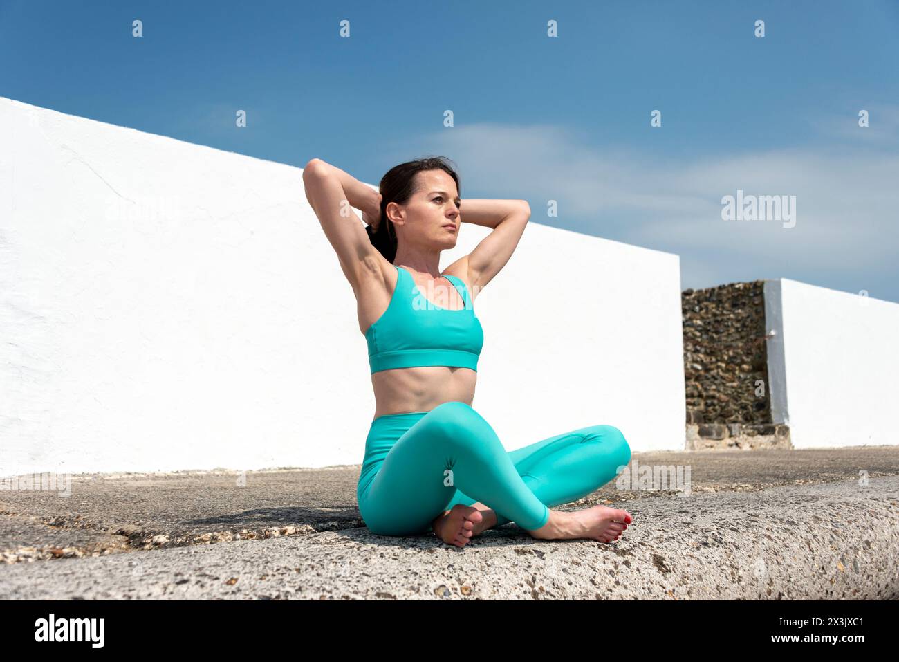 Sportliche Frau, die mit den Händen hinter dem Kopf sitzt und sich draußen in der Sonne ausdehnt Stockfoto